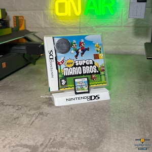 Pode incluir: Uma caixa de jogo Nintendo DS New Super Mario Bros. e um cartucho exibidos num suporte de plástico branco. A caixa apresenta o Mario e o Luigi a saltar, uma planta Piranha e um Koopa Troopa. O cartucho está posicionado à frente.