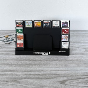 May include: A black plastic Nintendo DS game cartridge storage stand featuring a central slot and multiple side compartments. The base displays the white Nintendo DS logo and the text @my3dprintz. Several game cartridges are inserted into the slots.