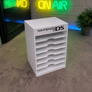 Könnte beinhalten: Ein weißer vertikaler Aufbewahrungsorganizer mit sieben horizontalen Regalböden und halbkreisförmigen Aussparungen für einfachen Zugriff. Die obere Vorderkante zeigt den schwarzen Aufdruck NINTENDO DS. Die Einheit hat einen soliden rechteckigen Rahmen.
