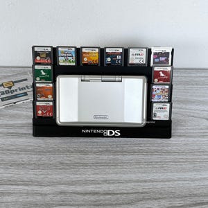 May include: A black plastic display stand for a Nintendo DS console and ten game cartridges. The stand features a central slot for the silver handheld console and side slots for the games. The text NINTENDO DS is printed in white on the base.