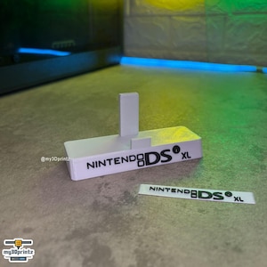 Può includere: Un espositore bianco stampato in 3D per console di gioco portatile con il testo nero NINTENDO DS XL sulla parte anteriore. Una targhetta rettangolare bianca separata con lo stesso testo nero NINTENDO DS XL è posizionata davanti al supporto.