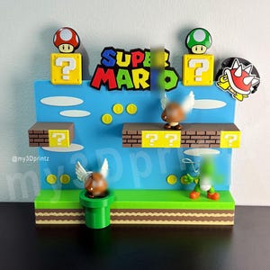 May include: A Super Mario-themed diorama featuring iconic elements from the video game. The scene includes a blue backdrop with the words "SUPER MARIO" in colorful letters, question mark blocks, mushrooms, coins, and a green pipe. A Goomba with wings is also present.