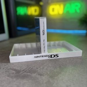 May include: A white plastic storage tray for Nintendo DS game cases, featuring the Nintendo DS logo printed on the front edge. A single game case titled New Super Mario Bros. is standing upright in the centre slot of the rectangular organiser.