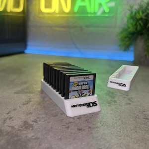 May include: A white plastic storage stand for Nintendo DS game cartridges, featuring the Nintendo DS logo on the front. The stand holds multiple black game cartridges, with one clearly labeled New Super Mario Bros. A second empty white stand is nearby.