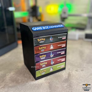 Op de afbeelding: Een zwarte 3D-geprinte opbergtoren met een blauw Game Boy Advance-logo aan de bovenzijde. De unit bevat vijf horizontale vakken, elk met een rechthoekige speldoos voor de Pokémon Emerald, FireRed, Sapphire, Ruby en LeafGreen versies.