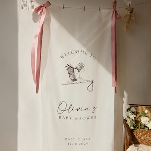 Op de afbeelding: Een crèmekleurige babyborrelbanner met de tekst "Welcome to Olivia's Baby Shower" en de datum 12.11.2025. De banner toont een illustratie van een ooievaar die een baby draagt. Roze linten en een teddybeer hangen boven de banner.