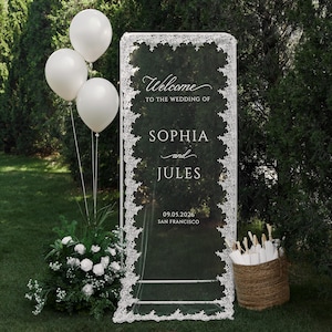 Op de afbeelding: Een hoog, rechthoekig welkomstbord voor een bruiloft van doorzichtig acryl met een witte kanten rand, voorzien van de tekst Welcome to the wedding of Sophia and Jules 09.05.2026 San Francisco, met drie witte ballonnen en een mand.