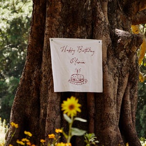 May include: A white banner with the words "Happy Birthday Olivia" in burgundy script is attached to a large tree. A drawing of a birthday cake is below the text. Yellow flowers are in the foreground.