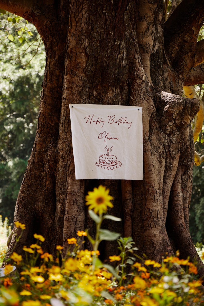 May include: A white banner with the words "Happy Birthday Olivia" in burgundy script is attached to a large tree. A drawing of a birthday cake is below the text. Yellow flowers are in the foreground.