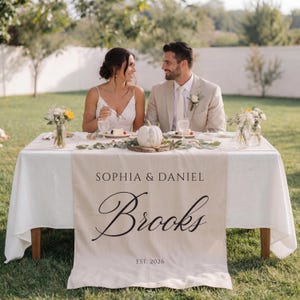 May include: A wedding table set for two, with a white tablecloth and a beige table runner. The runner is printed with the names "Sophia & Daniel" and the surname "Brooks" in script, with "EST. 2026" underneath. The table is decorated with flowers and a small pumpkin.