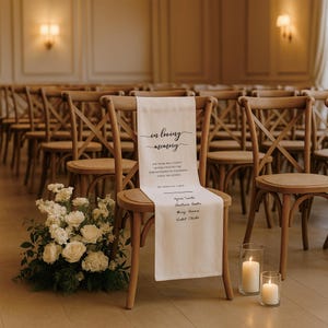 May include: A cream-colored chair sash with the words "in loving memory" in elegant script. The sash is draped over a wooden chair in a room with rows of similar chairs. A floral arrangement and lit candles are in the foreground.