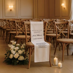 May include: A wedding ceremony scene with wooden chairs, a floral arrangement, and candles. A chair is draped with a white banner that reads "RESERVED in loving memory of James Whitmore FATHER OF THE GROOM."