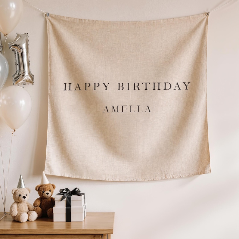 May include: A beige fabric banner with the words "HAPPY BIRTHDAY AMELLA" in black. The banner is hanging on a wall, with balloons and teddy bears in front of it. There are two gift boxes with black ribbons.