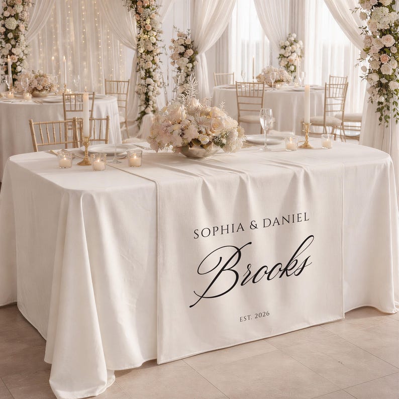 May include: A white tablecloth featuring the names "Sophia & Daniel" and the surname "Brooks" in black script, with "EST. 2026" below. The table is set for a wedding reception, with floral arrangements and candles.