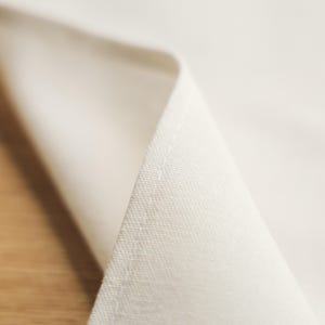 May include: Close-up of a folded, cream-coloured linen napkin. The fabric has a subtle texture and a visible seam along the edge. The napkin is folded to reveal a triangular shape, with the corner of the fabric pointing towards the viewer.