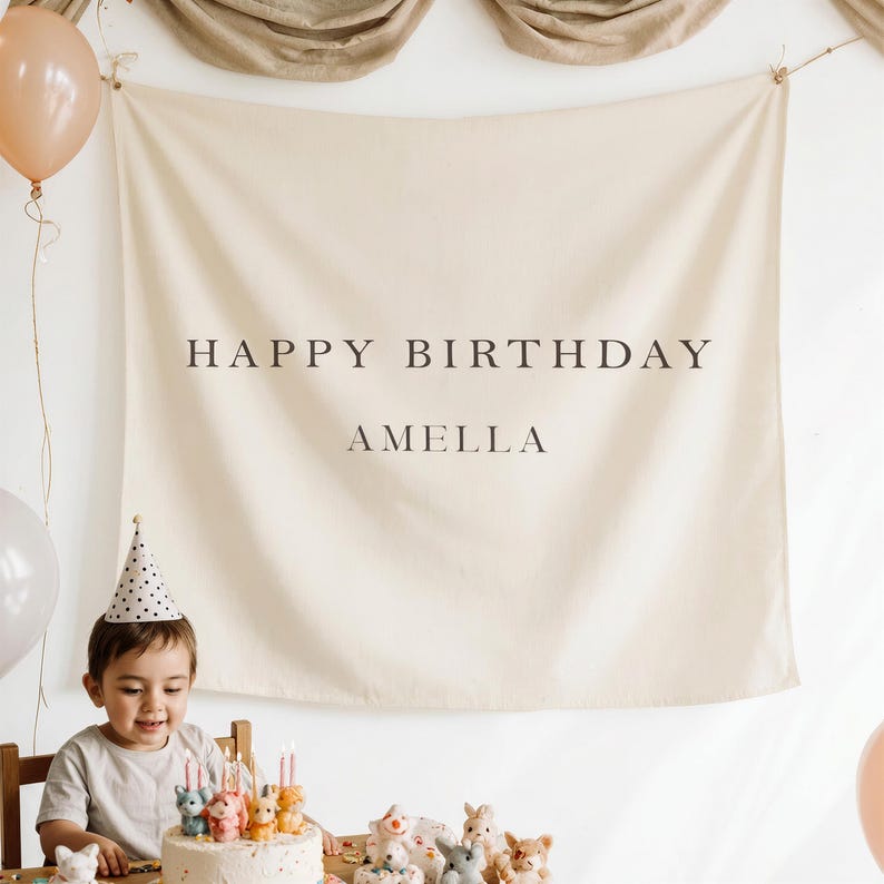 May include: A beige banner with the words "HAPPY BIRTHDAY AMELLA" in black lettering hangs on a wall. A young child wearing a party hat sits at a table with a birthday cake and balloons. The cake is decorated with candles and animal figurines.