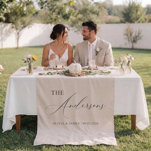 May include: A wedding table setting for two, with a white tablecloth and a beige table runner. The runner is printed with "The Andersons" and the date "09.05.2026". The table is decorated with flowers and a small pumpkin.