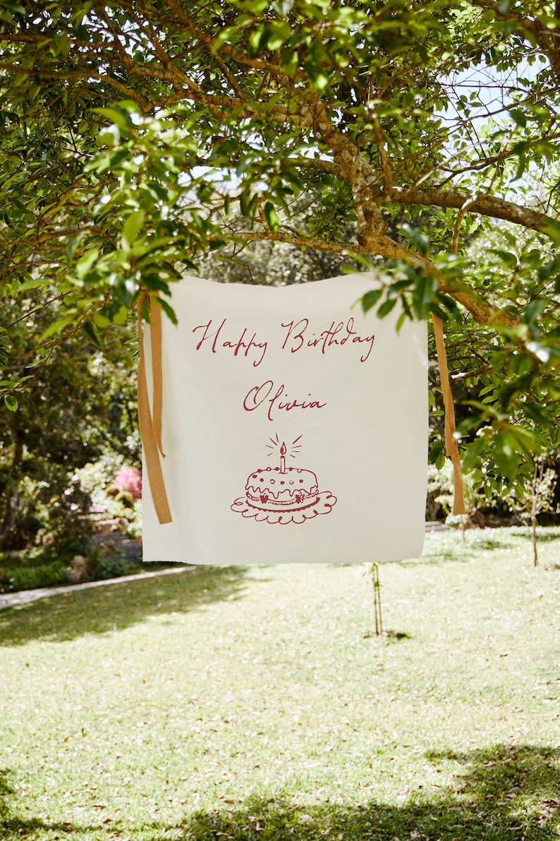 May include: A white banner with the words "Happy Birthday Olivia" in burgundy script, along with a cake illustration. The banner is hanging from a tree with brown ribbons. The scene is outdoors on a sunny day.
