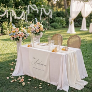 May include: A wedding reception table set outdoors with a white tablecloth, floral displays, and two wicker chairs. The table runner reads "The Andersons" with the wedding date. Tall glass candle holders and lemons are on the table.