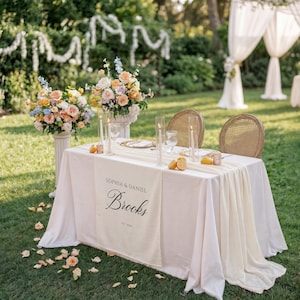 May include: An outdoor wedding reception table with a white tablecloth and floral arrangements. The table runner reads "Sophia & Daniel Brooks." Two wicker chairs face the table. Tall candles and lemons decorate the table.