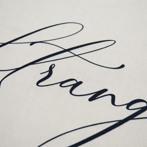 May include: A close-up shot of a cream-coloured fabric featuring the word "Arrange" in dark blue cursive. The elegant script has flowing lines, creating a sophisticated and stylish appearance.