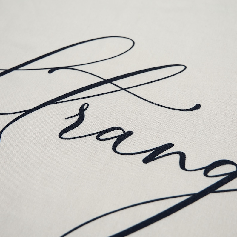 May include: A close-up shot of a cream-coloured fabric featuring the word "Arrange" in dark blue cursive. The elegant script has flowing lines, creating a sophisticated and stylish appearance.