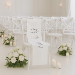 May include: A white chair with a white fabric runner that reads "In Loving Memory." The runner lists names and a message. White roses and candles decorate the aisle. The setting is a wedding ceremony.