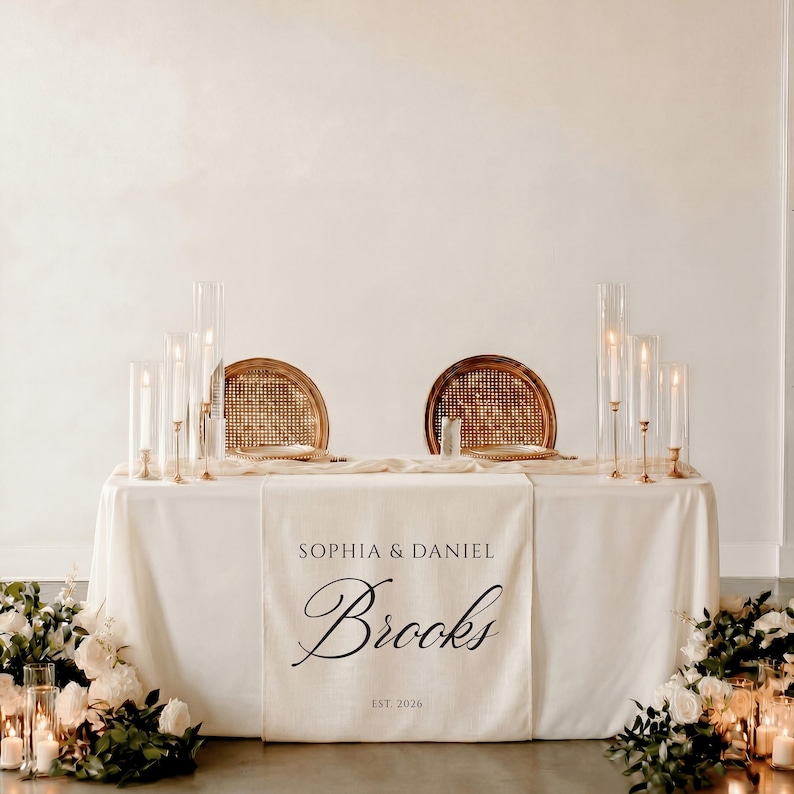 May include: A wedding table setting features a white tablecloth, two chairs with woven backs, and glass candle holders. A banner displays the names "Sophia & Daniel Brooks" and "EST. 2026". Floral arrangements and candles decorate the scene.