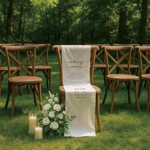 May include: A wooden chair with a white fabric runner that reads "in loving memory." The runner lists names and a message. White flowers and candles sit in front of the chair. Other wooden chairs are in the background.