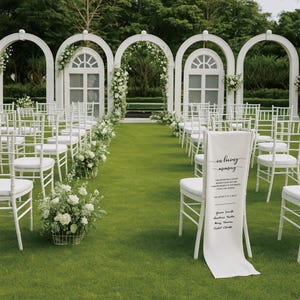 May include: An outdoor wedding ceremony setup with white chairs, floral arrangements, and decorative arches. A banner on a chair reads "in loving memory" with names and a date. The scene is set on a green lawn.
