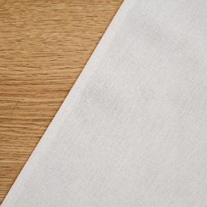 May include: A close-up of white linen fabric against a wooden background. The fabric is a pale, neutral shade, and the wood has a warm, natural grain. The image is well-lit, showing the textures of the fabric and wood.