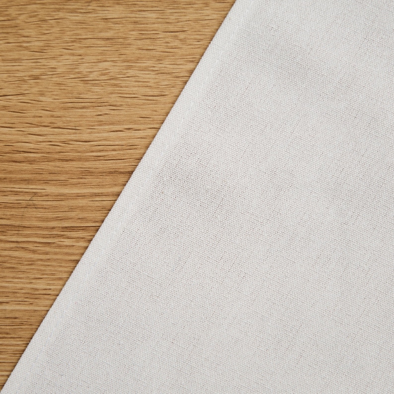 May include: A close-up of white linen fabric against a wooden background. The fabric is a pale, neutral shade, and the wood has a warm, natural grain. The image is well-lit, showing the textures of the fabric and wood.