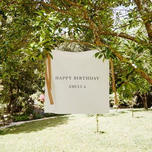 May include: A white rectangular banner with the text "HAPPY BIRTHDAY AMELLA" in black, hanging from a tree branch. The banner is secured with brown ribbons. The background features a green lawn and trees.