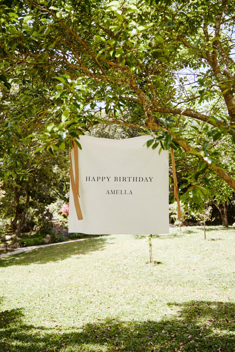 May include: A white rectangular banner with the text "HAPPY BIRTHDAY AMELLA" in black, hanging from a tree branch. The banner is secured with brown ribbons. The background features a green lawn and trees.