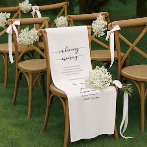 May include: Wooden chairs with white fabric draped over the backs, adorned with floral arrangements and ribbons. The fabric reads "in loving memory" with names listed below. A touching memorial for a wedding or event.