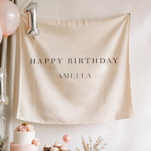 May include: A beige fabric banner with the words "HAPPY BIRTHDAY AMELLA" in black. A silver number one balloon and two pink balloons are visible. A two-tiered pink cake with flowers is in the foreground.