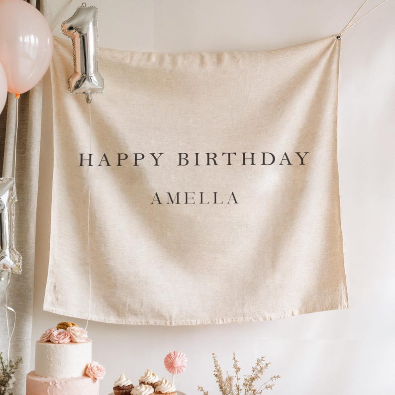 May include: A beige fabric banner with the words "HAPPY BIRTHDAY AMELLA" in black. A silver number one balloon and two pink balloons are visible. A two-tiered pink cake with flowers is in the foreground.