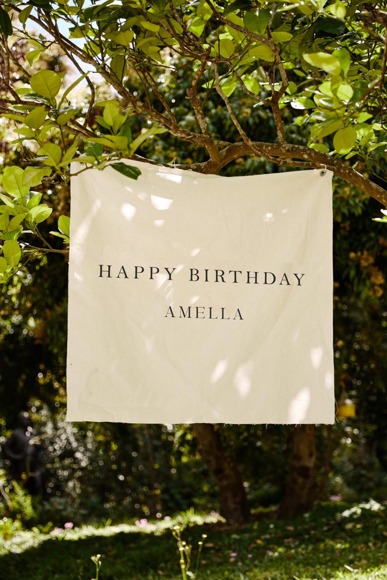 May include: A cream-colored banner with the words "HAPPY BIRTHDAY AMELLA" in black lettering hangs from a tree branch. The banner is rectangular and appears to be made of fabric. The background is a blurred view of green foliage and sunlight.