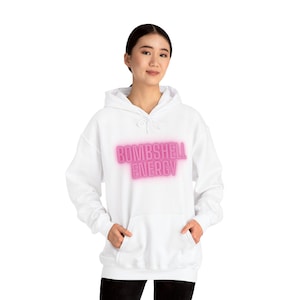 May include: White hooded sweatshirt with the text "BOMBSHELL ENERGY" in a pink, glowing font. The sweatshirt has a front pocket and a drawstring hood. The model is wearing the sweatshirt.