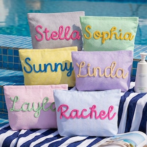 Könnte beinhalten: Eine Auswahl an pastellfarbenen Kosmetiktaschen mit Streifenmuster, jede mit einem Namen in Schreibschrift bestickt. Die Taschen werden in der Nähe eines Schwimmbads präsentiert, mit einer Zeitschrift und einem Handtuch im Vordergrund.