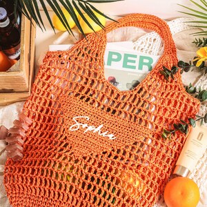 Puede incluir: Bolsa de mano de crochet naranja con el nombre "Sophia" escrito en blanco en la parte delantera. La bolsa est&aacute; hecha de un material grueso y duradero y tiene una parte superior ancha y abierta.