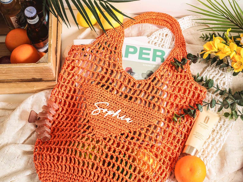 Puede incluir: Bolsa de mano de crochet naranja con el nombre "Sophia" escrito en blanco en la parte delantera. La bolsa est&aacute; hecha de un material grueso y duradero y tiene una parte superior ancha y abierta.