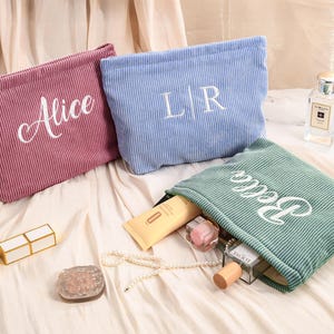 Peut inclure: Trois trousses de maquillage en velours c&ocirc;tel&eacute; rose, bleu et vert, chacune brod&eacute;e d'un nom ou d'initiales en blanc. Le sac vert est ouvert, r&eacute;v&eacute;lant des produits de beaut&eacute;. Un rouge &agrave; l&egrave;vres, un pinceau et un flacon de parfum sont &eacute;galement visibles.
