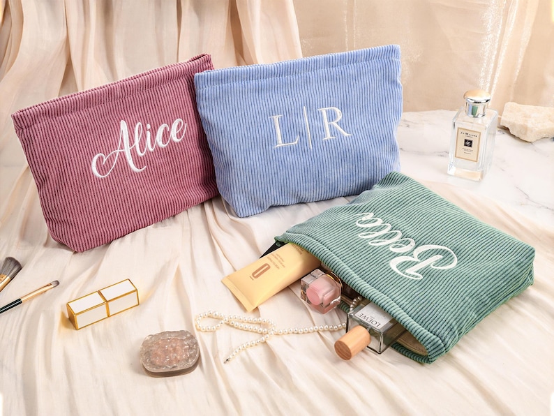 Peut inclure: Trois trousses de maquillage en velours c&ocirc;tel&eacute; rose, bleu et vert, chacune brod&eacute;e d'un nom ou d'initiales en blanc. Le sac vert est ouvert, r&eacute;v&eacute;lant des produits de beaut&eacute;. Un rouge &agrave; l&egrave;vres, un pinceau et un flacon de parfum sont &eacute;galement visibles.