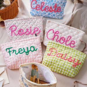 Può includere: Una collezione di pochette in tessuto trapuntato con vari motivi floreali e a quadretti, ognuna con un nome ricamato in rilievo con filo. Le pochette sono blu, rosa, bianche e verdi, una è aperta e mostra dei pennelli da trucco.