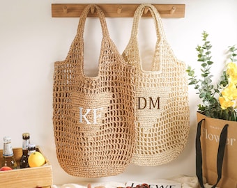 Op de afbeelding: Twee beige gehaakte tassen hangen aan haken. De tas aan de linkerkant heeft de initialen "KF" geborduurd. De tas aan de rechterkant heeft de initialen "DM" geborduurd.