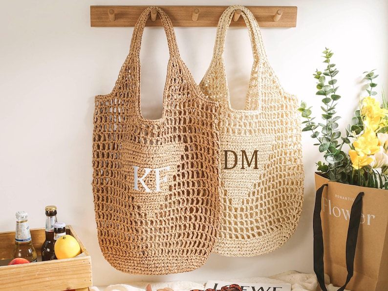 Puede incluir: Dos bolsas de mano de crochet beige colgadas de ganchos. La bolsa de la izquierda tiene las iniciales "KF" bordadas. La bolsa de la derecha tiene las iniciales "DM" bordadas.