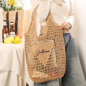 Puede incluir: Una bolsa de mano de crochet de color tostado con la palabra "Victoria" bordada en la parte delantera. La bolsa la lleva una persona que lleva una camisa blanca y unos vaqueros azules.