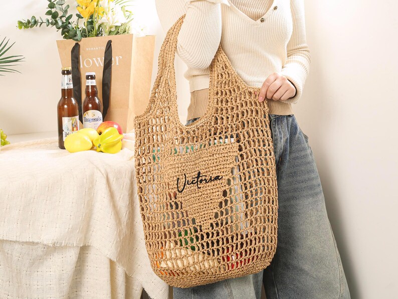 Puede incluir: Una bolsa de mano de crochet de color tostado con la palabra "Victoria" bordada en la parte delantera. La bolsa la lleva una persona que lleva una camisa blanca y unos vaqueros azules.