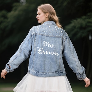 May include: A light blue denim jacket with pearl embellishments on the back. The jacket has the text "Mrs. Brown" written in white on the back.